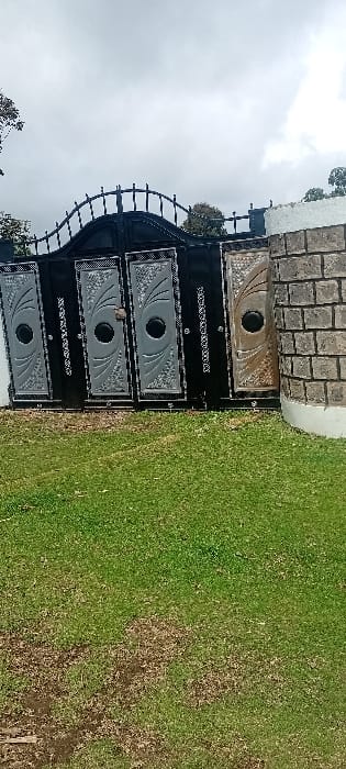 The scene depicts the exterior of a property in the city of Rwanyambo, Kenya, under an overcast sky. A closed, ornate metal gate dominates the midground, featuring a black frame with an arched, decorative top section composed of curved vertical bars. Below, four distinct rectangular panels are arranged horizontally. The two leftmost panels are silver-grey with intricate swirling patterns and a central black circular motif. The third panel from the left is black with the word "MARANATHA" inscribed vertically in white text. The rightmost panel is gold-bronze with a similar swirling design and central black circle. To the immediate right of the gate, a stone pillar constructed from stacked grey blocks and topped with a white cap is visible. The foreground consists of green grass with scattered brown patches. Trees are present in the background, visible above and behind the gate. No people or animals are observed.