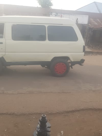 The image displays an outdoor scene in Kufena, Nigeria, featuring a white commercial-style van parked on a dusty, unpaved surface. The van, positioned on the left and central parts of the frame, shows its passenger side with multiple dark-tinted rectangular windows.
Towards the rear of the van, a single wheel with a black tire and a distinct red center cap is visible on the ground. This wheel appears to be mounted on a dark frame or stand, suggesting it might be a spare or part of a separate piece of equipment. Further to the right, resting on the dusty ground, is a dark metallic engine component resembling a crankshaft, identifiable by its lobed structure and a geared end.
In the background, behind the van, stands a building with a corrugated metal roof and what appears to be brick or mud walls. A white rectangular sign is mounted on this building, bearing partially legible text: "DAYLAB CARPENTRY & SERVICES NIG. LTD." The scene is brightly lit, indicating it is daytime. No people are visible, and no specific activity is occurring; the van is stationary, and the various mechanical parts are simply lying on the earthy ground.