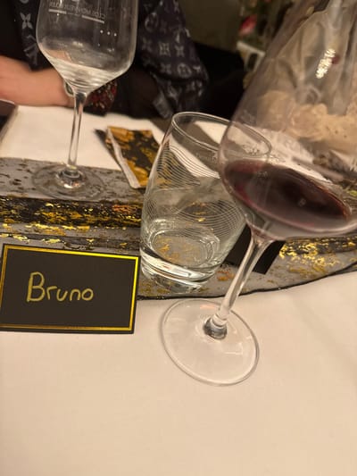 An overhead shot displays a table setting, dominated by glassware. A red wine glass spills into a water glass adorned with subtle patterns. A second, empty wine glass sits nearby. A black place card with gold trim and the name "Bruno" in gold lettering is positioned beside the water glass. The table runner features black lace with gold foil accents. A white tablecloth provides a clean backdrop. A Louis Vuitton bag is faintly visible in the background.