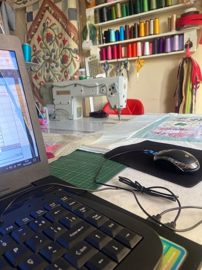 The image depicts a sewing room, likely a home studio, in Vila Velha, Brazil. The scene is a close-up view from the perspective of someone sitting at a table, with a laptop in the foreground. A sewing machine, identified by the "Singer" logo, is positioned on a large white table behind the laptop. The table also has fabric laid out, suggesting ongoing sewing projects, and a quilted wall hanging is visible in the background. Shelves above are filled with spools of colorful thread and various sewing accessories like scissors. A black computer mouse rests on a mousepad to the right of the laptop, with its cord neatly tied. The overall impression is one of a dedicated workspace for crafting and sewing.