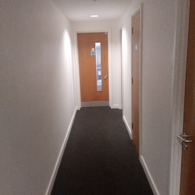 The image depicts an indoor hallway setting, likely within a commercial or residential building in Coventry, United Kingdom. The corridor features light-colored walls and a dark gray carpet. Two visible doors, one at the end of the hallway and another on the right, are made of light brown wood. The door at the end of the hall has a vertical window and some signage, including an emergency exit sign. The hallway is illuminated by overhead lighting. No people are visible in the scene.