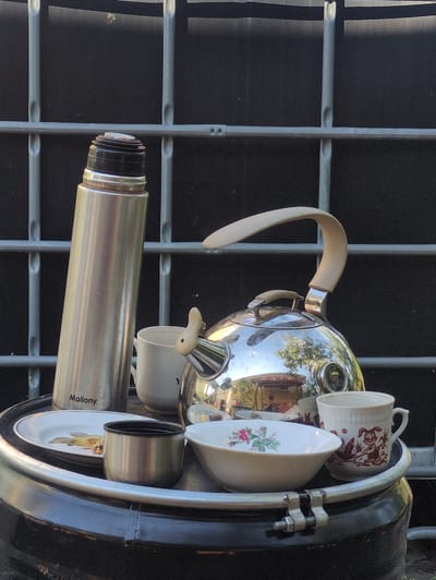 This image shows a collection of vintage-style kitchenware, including a chrome kettle with a cream-colored handle, two ceramic cups with decorative patterns, and a bowl with a floral design, all placed inside a black, cylindrical object that appears to be a barrel or a large container. A silver thermos is positioned in front of the kettle. The items are arranged on a surface with a grid-like pattern, possibly a metal grate or fencing, against a dark background. Reflections on the chrome kettle suggest an outdoor setting with trees and buildings visible. The lighting indicates it is daytime, likely sunny. The word "Mallory" is visible on the thermos. The scene appears to be set up as a display or arrangement of these items, possibly in a garden or an outdoor market setting in Velikovo, Russia.