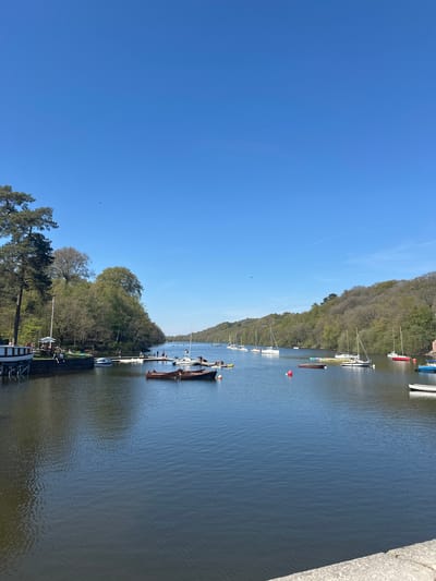The image depicts a scenic view of a body of water, likely a river or lake, in Staffordshire Moorlands, United Kingdom. A line of boats and yachts of varying sizes is docked along the water's edge, indicating a marina or waterfront area. The opposite side of the waterway is lined with a dense forest, and the sky is a clear, bright blue. In the lower portion of the image, a path or walkway can be observed alongside the water, with people walking or sitting on benches. The overall atmosphere is calm and natural, suggesting a recreational area.