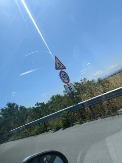 A road sign is mounted on a pole against a bright blue sky with a strong sun glare reflecting off a windshield. The sign displays three symbols: a triangular warning sign with a slippery road icon, a circular sign with the number "90", and a square sign with a snowflake symbol. Beyond the sign, a metal guardrail runs along the side of a paved road, bordered by dense green and dry brush vegetation. In the distance, rolling hills and fields are visible under the clear sky. The image appears to be taken from a moving vehicle, as evidenced by the reflection in what seems to be a car's side mirror in the lower foreground. The location is identified as Mirovo, Bulgaria.