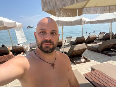 The image is a selfie taken at a beach in the city of Pallini Municipal Unit, Greece. The foreground features a man with a shaved head and a beard, wearing a black cord necklace with a wooden cross pendant. He is shirtless and appears to be wet, possibly from swimming. Behind him, a sandy beach is lined with rows of unoccupied lounge chairs and umbrellas. The sea is visible in the background with a few people swimming and a boat anchored offshore. The sky is clear and blue, indicating a sunny day. The overall scene depicts a relaxing day at a beach resort.