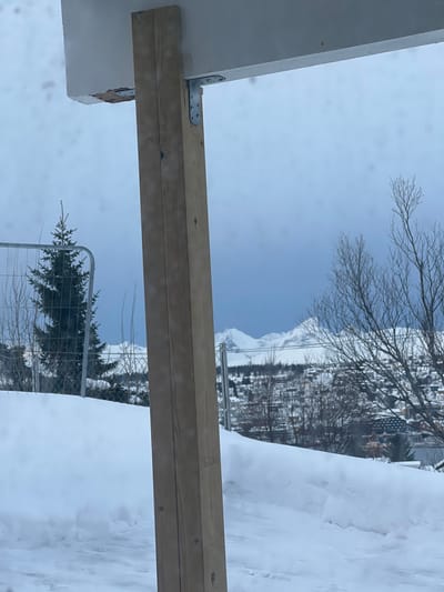 The image displays a static outdoor winter scene in Tromsø, Norway. In the immediate foreground, a vertical, light-colored wooden post, constructed from two timber planks, rises centrally, partially obscuring the view. This post is secured to a horizontal, white-painted overhead beam by two silver-colored, L-shaped metal brackets fastened with screws; one bracket is visible on the left, the other on the right. The underside of the beam near the left bracket shows minor structural damage or unfinished material. The ground level below is covered in a thick, undisturbed layer of white snow.

The midground features a prominent snowdrift or bank, behind which a chain-link fence is visible on the left, partially obscuring a conical evergreen tree. Scattered bare deciduous trees and sparse bushes are also present. Further in the midground, the snow-covered rooftops and structures of a dense urban area, consistent with the city of Tromsø, descend down a hillside, intermingled with more bare trees.

The background is dominated by rugged, snow-capped mountains with defined peaks under an overcast, light blue-grey sky. No individuals or animate subjects are present in the scene.