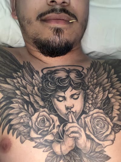 The image shows a close-up view of a person's chest with a tattoo of an angel surrounded by roses. The person has facial hair, including a mustache and goatee, and a toothpick is visible in their mouth. The tattoo is black and gray and covers a significant portion of the chest. The background is a white sheet or pillow. The location of Milton Keynes, United Kingdom, is not depicted in this image.