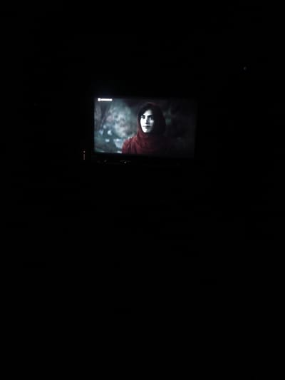 The image displays a television screen in a very dark room, suggesting it is nighttime or a room with minimal external light. On the television screen, a still image of a young woman is visible. She has dark hair and is wearing a dark red or maroon hooded garment that covers her head and shoulders. Her gaze is directed slightly away from the camera, and her expression appears neutral or contemplative. The background behind the woman on the screen is blurred, showing shades of green and grey, indicative of an outdoor, possibly wooded setting.

In the upper-left corner of the television screen, a small, light-colored logo or text is partially visible; it seems to read "ASTRONAU" or a similar fragment. Below the television, a media device is present, indicated by a series of small, glowing indicator lights—some amber and some green—suggesting it is powered on. The immediate surroundings of the television and device are obscured by profound darkness. The overall scene depicts the activity of watching television in a dimly lit environment. The location context provided is Stroevo, Bulgaria, although no specific visual cues within the image confirm this location.