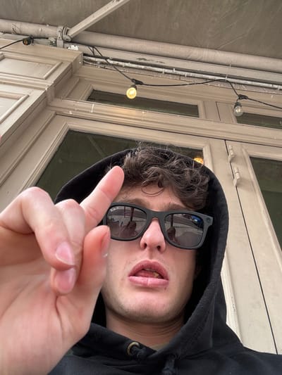 The image captures a male individual in a close-up, selfie-style shot taken outdoors in Vincennes, France. The subject, a young adult with short, dark, curly hair, wears a black hooded sweatshirt with the hood pulled up. He is wearing black rectangular Ray-Ban sunglasses, which reflect indistinct figures and an outdoor scene. His mouth is slightly agape, revealing teeth and a portion of his tongue, on which a small, light-colored particle is visible. His left hand is raised into the foreground, with the index finger extended vertically. The background consists of a light-colored building facade featuring paneled architecture and large windows. Above the facade, a light-grey awning extends. String lights with illuminated, warm-toned bulbs are strung horizontally across the upper part of the frame, attached to the building structure.