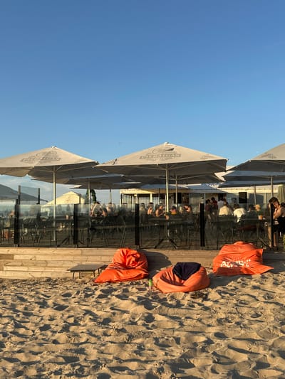 The image depicts an outdoor seating area on a sandy beach, likely a cafe or restaurant, in Jūrmala, Latvia. The scene is set during the day, indicated by the bright sunlight and clear blue sky. The perspective is from the sandy ground looking upwards towards the establishment and the sky.
The foreground is dominated by sand, with several large, bright orange beanbag chairs or cushions scattered on it. One of these cushions has dark blue fabric peeking out from beneath it, and another has a white logo or text printed on it. A green plastic bottle is also visible on the sand.
Behind the sand is a wooden deck, which forms the floor of the seating area. The seating area itself is partially covered by large, light-colored umbrellas, some of which have logos or text printed on them. Through the glass walls of the establishment, several patrons can be seen sitting at tables, engaged in conversation or enjoying their time. The presence of umbrellas and patrons suggests a lively atmosphere, likely a popular spot for relaxation and dining. The building appears to have multiple levels of seating.
The overall mood is relaxed and summery, consistent with a beachside location. The time of day appears to be late afternoon or early evening, as evidenced by the long shadows cast by the beanbag chairs and the warm sunlight.