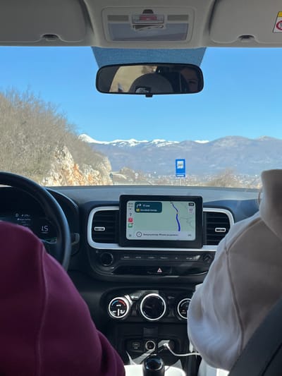 The image depicts the interior of a vehicle, viewed from a rear passenger seat, looking forward through the windshield. The scene is located in Miločani, Montenegro.

Two occupants are partially visible. On the left, the driver's left arm and shoulder are visible, clad in a dark maroon long-sleeved garment. On the right, the front passenger's right arm and shoulder are visible, wearing a light-colored hoodie. The front passenger's upper head and face are reflected in the rearview mirror, revealing a grey beanie with a "THE NORTH FACE" logo.

The vehicle's black dashboard is prominent, featuring a central infotainment screen displaying a navigation map. The map shows a route with "11 KM" remaining to a destination identified as "Народних Нареја" (Narodnih Nareja). The screen also indicates the time as 10:43 and a message stating "Аккумулятор iPhone разряжен" (iPhone battery discharged). Below the screen are climate controls, and further down, two USB-A ports are visible, one with a white charging cable plugged in. A black steering wheel is partially visible on the left, with a digital display behind it showing "D" and "1.5". An overhead console above the windshield contains interior lights and an SOS button.

The exterior environment features a clear, bright blue sky. On the immediate left, a rocky hillside with sparse, bare deciduous trees is visible. In the distance, prominent snow-capped mountains form the horizon. A paved road is visible, bordered by a guardrail on the right side of the frame below the mountains. A blue rectangular road sign, depicting a bus icon, is located on the right side of the road in the mid-distance, indicating a bus stop. A cluster of buildings, consistent with a town or settlement, is discernible in the valley beneath the distant mountains. The vehicle is in motion, evidenced by the active navigation system and the 'D' indicator on the dashboard display.