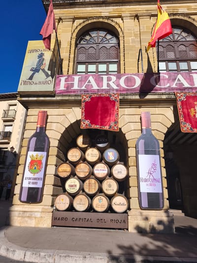 The image depicts a festive outdoor scene in the city of Haro, Spain, likely during a wine-related event. In the foreground, a large, stylized advertisement of a wine bottle stands to the left, featuring a label with the "Ayuntamiento de Haro" (City Council of Haro) emblem and text. To its right, a pyramid of wine barrels is displayed, each bearing different winery labels and names such as "ARVS," "BODEGAS," "RODA," "MUGAS," "AKUTAIN," "MANZANOS," and "SANTALBA." Above the barrels, two large banners are hung, one with the text "HARO CAPITAL" and another featuring the Spanish flag and red flags. The background consists of a building with arched windows and stone architecture. Shadows suggest it is a sunny day. Two women are visible in the background on the left side of the image, near a building with balconies.