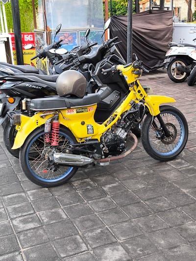 The image depicts a row of parked motorbikes in the city of Tibubeneng, Indonesia.  In the foreground is a bright yellow motorcycle, custom built and heavily modified, with blue rims.  Behind it are two modern black motorbikes, one with a brown helmet resting on the seat.  To the right is a white and brown motorbike with a license plate.  In the background are advertisements and a brown tarp structure. The motorbikes are parked on a paved area with square stones. There is also some signage on a small white structure.