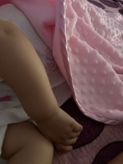 A close-up shot captures a baby's leg and foot resting on a pink textured blanket and a white surface. The baby's skin is smooth and fair. The pink blanket has a pattern of raised dots, creating a soft, plush texture. The white surface beneath appears to be fabric, possibly clothing or another blanket, with faint floral patterns visible. The lighting is soft, suggesting an indoor setting. There are no discernible elements in the image that indicate a specific location within Parvomay, Bulgaria.
