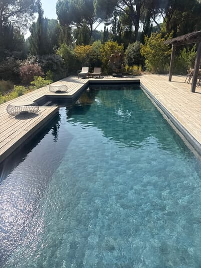 This image depicts a private swimming pool area in Grândola, Portugal. The scene is outdoors, with a focus on a rectangular swimming pool with turquoise-tiled lining, surrounded by a wooden deck. The pool is illuminated by natural sunlight, casting reflections on its surface. Two black wire mesh tables with intricate designs are placed on the wooden deck. In the background, lush greenery and trees are visible, creating a natural and serene environment. To the right of the pool, a wooden pergola with a thatched roof shelters a dining table and chairs. There are no people visible in the image. The overall atmosphere is tranquil and inviting, suggesting a leisure space.