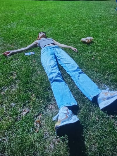 A female individual lies supine on an expanse of green grass in Kasagaran, Uzbekistan. She is dressed in a light grey tank top, light blue jeans, and white athletic shoes with tan and yellow accents. Dark sunglasses cover her eyes, and reddish-brown hair is fanned out around her head. Both arms are extended horizontally to her sides with palms facing upwards, and her legs are fully extended. A multi-colored rectangular object, consistent with a packet or small device, rests on the grass near her left hand. To the right of her torso, a crumpled light-brown object, appearing to be paper or fabric, is visible on the grass. Sparse brown leaves are intermingled with the grass. The scene is illuminated by direct sunlight, casting distinct shadows.
