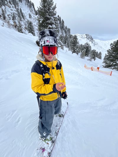 A snowboarder in a yellow and black jacket, grey pants, and red goggles sits in the snow. A black beanie covered in snow sits atop their head. Behind, trees covered in snow line the mountainside, and a safety fence runs alongside the slope. The sky is overcast. The snowboarder is centered in the frame and faces the viewer.