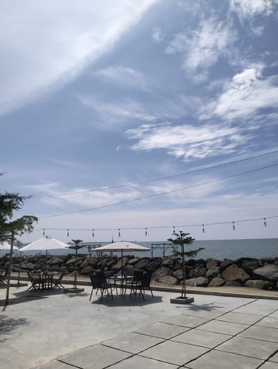 This media file depicts an outdoor seating area with a view of the ocean, likely a cafe or restaurant. The scene is set under a bright, partly cloudy sky, suggesting a pleasant day. The time appears to be daytime, possibly late morning or early afternoon, due to the clear visibility and the way the light falls.
In the foreground, a paved area with square concrete tiles serves as the seating space. Several tables and chairs are arranged, some covered by white and light brown umbrellas, providing shade. A few small trees are planted in concrete bases, adding a touch of greenery to the area. String lights with exposed bulbs are strung across the scene, hinting at possible evening use.
Beyond the seating area, a rocky seawall separates the paved space from the ocean. The water appears calm, with a gentle rippling surface. In the distance, the horizon is visible, with some hints of structures or ships. The overall setting suggests a tranquil coastal location, perhaps a tourist spot or a relaxed dining establishment. The image conveys a sense of peacefulness and openness, inviting viewers to imagine themselves enjoying a meal or a drink by the sea. No people are visible in the image. The location context places this scene in Lancok, Indonesia.