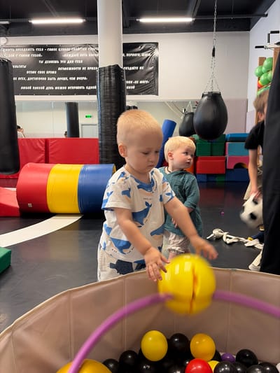 This image captures a scene inside a children's sports or play facility in Moskovskiy, Russia. Two young Caucasian boys are the primary subjects. The boy in the foreground, facing right, is wearing a white t-shirt with a blue whale print and white shorts. He is reaching into a large, light-colored playpen filled with black and yellow balls, with a blurred yellow ball in his hands and a purple hoop visible. To his left and slightly behind him, a second boy with blonde hair, wearing a teal long-sleeved shirt and striped shorts, looks towards the right, seemingly observing something out of frame.
The environment suggests an indoor active play area. A large, cylindrical mat with red, yellow, and blue sections lies on the floor behind the boys. In the background, punching bags are visible, one hanging from the ceiling. Colorful building blocks are stacked on shelves. A large banner with text in Russian is mounted on the wall above. The floor is dark, possibly a gym mat. The lighting appears to be overhead industrial fluorescent lights, characteristic of such facilities. A partial view of an adult, possibly supervising, is visible on the far right.