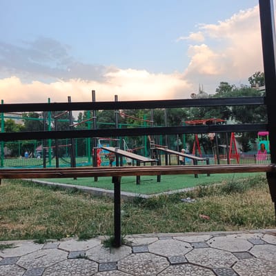 The image shows an outdoor playground at dusk. The sky is a pale blue with soft, fluffy clouds that are tinged with pink and orange from the setting sun. A dark metal fence runs across the foreground, partially obscuring the view of the playground.
The playground itself features a variety of equipment, including monkey bars, swings, and climbing structures. Some of the equipment is made of metal and painted in bright colors like red and green, while other parts are made of wood. The ground is covered with artificial turf, giving it a bright green appearance. In the background, there are trees and buildings, suggesting an urban or suburban setting.
Several people are visible in the playground. In the middle ground, a few children are playing on the climbing equipment, with one child hanging from the monkey bars and another appearing to be on a seesaw. In the background, two figures, likely children, are playing soccer on a fenced-in field. A young boy in a white cap stands near a seesaw in the right middle ground.
The overall mood of the image is peaceful and relaxed, characteristic of an early evening at a public park. The time of day is indicated by the soft lighting and the colors of the sky. The location context suggests Angren, Uzbekistan. There is no visible text in the image.