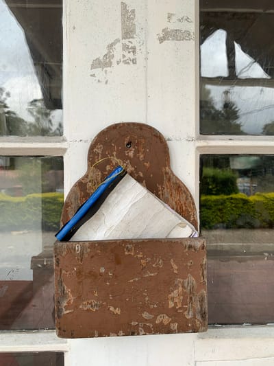 In Nuwara Eliya, Sri Lanka, a weathered, brown wooden letter holder is mounted on a white wall next to a window with panes of glass. The letter holder contains a blue folder with a white object inside. The white wall exhibits peeling paint, revealing patches of a lighter material underneath. The window glass is somewhat grimy and reflects the surrounding environment, which includes greenery and cloudy skies. The overall scene suggests a rustic or older building, possibly a colonial-era structure common in Nuwara Eliya.