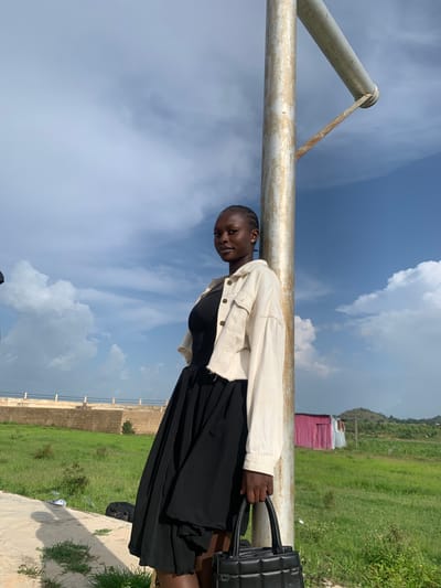 A young Black woman stands leaning against a rusty metal pole in Gwafan, Nigeria. She is wearing a black dress and a cream-colored jacket, holding a black handbag. The background shows a grassy field with a brick wall and a small pink building. The sky is cloudy.