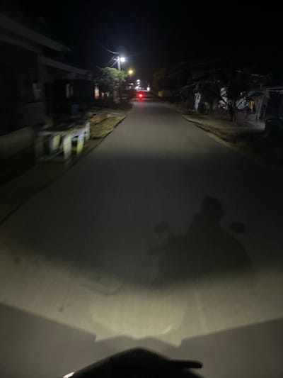 A nighttime shot reveals a street lined with buildings and parked vehicles. Streetlights illuminate the scene, casting shadows along the sidewalk. A red light is visible, potentially indicating a traffic signal. The photo is oriented sideways, showcasing the street vertically with the dark night sky and ground on either side. A blurry patch obscures the right side of the image.