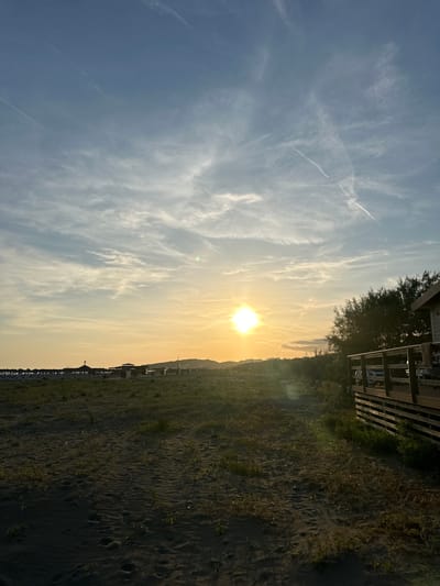 The image shows a beach scene in Donji Štoj, Montenegro, at sunset. The sun is positioned near the horizon, casting a bright golden glow across the sky and the beach. Wispy clouds are scattered throughout the sky, with contrails from airplanes visible. The beach appears to be composed of dark sand with sparse vegetation. In the distance, there are structures along the beach, likely beach umbrellas and small buildings. A wooden deck structure is partially visible at the top right of the image.