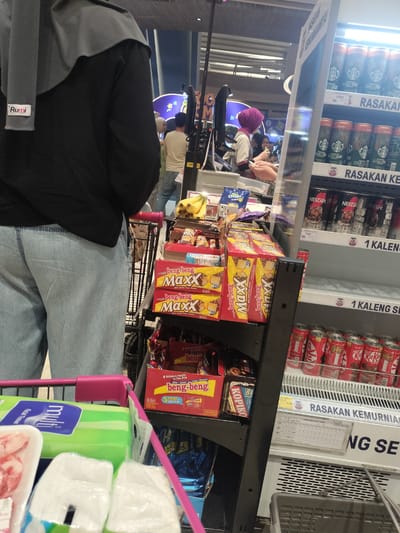 In a supermarket aisle in Gunung Putri, Indonesia, a person wearing a black long-sleeved shirt and light blue jeans is pushing a shopping cart. The cart contains a package of Multi Facial Tissues, a package of raw meat, and several boxes of Beng-Beng Maxx chocolate bars. To the right of the cart, a refrigerated display case is filled with various canned beverages, including Starbucks Double Shot Espresso and Kit Kat. Some shelves are empty, while others hold cans of coffee. A price tag for "NESCAFE ICE BLACK" is visible on one shelf. The background shows other shoppers and store displays.
