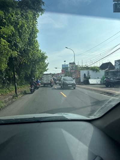 Captured from inside a vehicle, the image presents an eastward view of a roadway in Jimbaran, Indonesia. The road, marked with yellow lines, accommodates several vehicles, including a dark-colored sedan directly ahead and a white truck further in the distance. Motorbikes, with riders wearing helmets, are seen weaving through traffic. Buildings and billboards line the sides of the road, with lush, green trees and foliage bordering the road's edge. Overhead, power lines extend across the sky. The sky is bright, indicating daytime. The vehicle's dashboard and a portion of the side window are visible in the foreground.