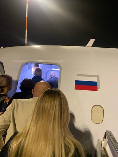 The image depicts a nighttime scene at Ben-Gurion Airport, Israel, showing individuals boarding a white aircraft. The perspective is from behind the queue of passengers ascending a set of airstairs, which are partially visible on the right.
The central subject is the side of a white airplane fuselage with its main cabin door open, revealing a brightly lit interior. A red, blue, and white horizontal striped flag, consistent with the flag of Russia, is affixed to the fuselage to the right of the open door. Below this flag, a small, oval-shaped aircraft window is visible. Inside the open door, a sign with "ВЫХОД EXIT" (Emergency Exit, in Cyrillic and English) is discernible. A bright light source, likely a pole-mounted floodlight, is visible in the upper-left, illuminating a striped pole (white and orange segments) above the aircraft.
Approximately six individuals are in the frame, primarily seen from behind as they queue to board the aircraft. In the immediate foreground, facing away, is a person with long, blonde hair. In front of them, a bald individual wearing a light-colored jacket is present. Further into the queue, a person with dark, curly hair is visible, followed by another individual in a dark jacket. Inside the aircraft's doorway, a person wearing a white shirt, possibly a crew member, stands facing the boarding passengers. Another individual, partially obscured by the person with dark hair, is also seen ascending into the cabin. The overall environment is dark, typical of an airport tarmac at night, with artificial lighting focused on the boarding area.