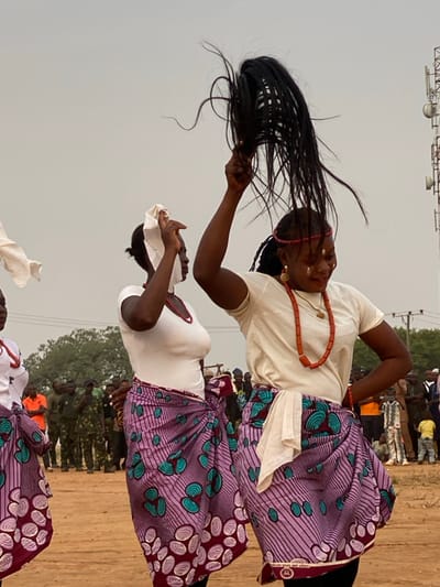 This dynamic image, taken in Agunu, Nigeria, depicts an outdoor cultural event featuring women dancing. In the foreground, a woman with dark skin is captured mid-movement, her long, dark braided hair dramatically flung upwards and to her left, held by her hand. She wears a plain white short-sleeved top, a striking red beaded necklace, and small gold earrings. Her vibrant wrap skirt, made of Ankara print fabric, displays patterns in purple, teal, and maroon/brown. A thin red band is visible on her head.
Behind her, another woman also dances, dressed in a similar white top and a matching patterned skirt. She holds a white cloth or head covering in her hands near her face. Both women are actively engaged in their performance, conveying energy and cultural expression.
The scene unfolds on dusty ground under an overcast sky, suggesting it is daytime. In the background, a crowd of people, some appearing to be spectators, are visible, along with distant trees and indistinct structures. The general ambiance points to a lively community gathering or festival. Faint text, possibly "ORIGINAL WAX PRINT," is discernible on the selvage edge of the fabric on the dancers' skirts.