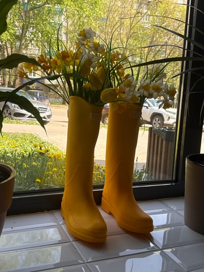 Two yellow rain boots filled with flowers are positioned on a window sill. The flowers include daffodils and tulips. Outside the window, a street scene with cars and yellow dandelions is visible. The location is Borok, Russia. The window sill is covered in white ceramic tiles.