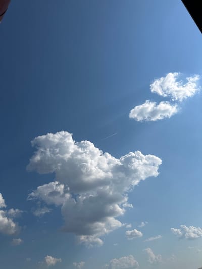 The image shows a clear blue sky with scattered white clouds. A thin, white contrail from an aircraft is visible diagonally across the upper right portion of the frame. The bottom edge of the image shows a dark, solid object, likely part of a structure or frame, obscuring the lower left corner. The scene is an outdoor view.