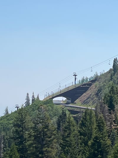 The image shows a ski lift system in a mountainous, forested area. The ski lift appears to be a gondola system, with multiple chairs suspended from cables running up a steep, tree-covered mountainside. The cables are supported by tall poles, and there are also what appear to be protective nets or barriers along the side of the mountain.

In the foreground, the bright blue sky dominates the left side of the frame. On the right side, dense green evergreen trees cover the slope. A dark, arched structure, possibly a tunnel or bridge, is visible partway up the mountain, with the ski lift cables passing over it. The overall scene suggests a sunny day, likely in the late spring, summer, or early fall, given the clear sky and lush greenery. The ski lift implies the location is a ski resort or a recreational mountain area. There is no visible text in the image. The image appears to be taken from a moving vehicle or from a lower elevation, looking up the mountainside.