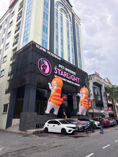 A vertical shot shows a modern veterinary medical center in Ampang Jaya Municipal Council, Malaysia. The building is beige with blue windows and a black lattice structure covering the entrance. "KLINIK VETERINAR" is displayed on the building. "PET UNIVERSE STARLIGHT VETERINARY MEDICAL CENTRE" is prominently displayed above the entrance. Inflatable orange and white cats adorn the front of the building. Cars and motorcycles are parked along the street in front of the building, and a person is standing near them. The sky is overcast.