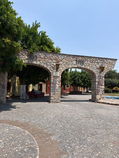 The image shows a stone archway in Cuautla, Mexico, built in 1938. The archway consists of two arches. The left side of the archway is partially covered by a tree. A cobblestone road leads through the archway. In the background, a building with red trim is visible. A person sits on a stone bench. The sky is clear.