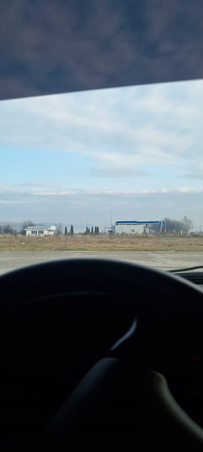 The image depicts an outdoor scene in Dragushinovo, Bulgaria, viewed from inside a vehicle. The bottom third of the frame is dominated by the dark, curved top portion of a steering wheel and a vehicle dashboard. Above this, the horizon reveals a flat landscape with sparse, light brown, dry vegetation. On the left, a white, single-story building is visible, featuring multiple rectangular windows and the Cyrillic text "ABTOMОЙКА" (Car Wash) on its facade. To the right of this building, a line of approximately seven slender, dark green coniferous trees stands. Further right and slightly beyond, a gas station structure with a prominent blue canopy and red digital price displays is identifiable. The sky above is partially cloudy, displaying patches of light blue amidst scattered white and gray clouds. No individuals are visible. The overall environment suggests a commercial or roadside area.