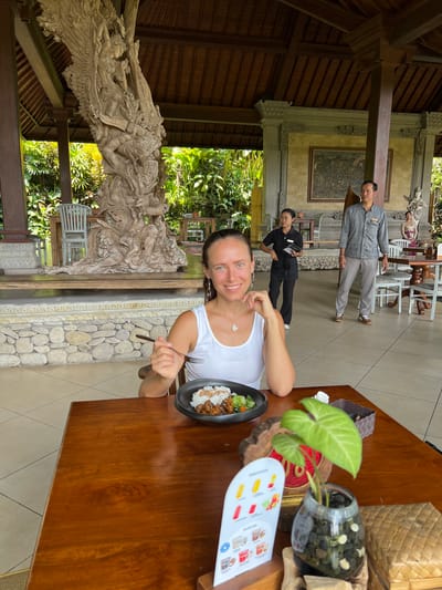 A woman sits at a wooden table in a restaurant in Kedewatan, Indonesia. She is wearing a white tank top and holds a fork. On the table is a plate of food, a plant in a glass vase filled with rocks, a menu, and a woven basket. A waiter and a waitress stand behind her. In the background is a detailed stone carving, lush greenery, and an open-air dining area.