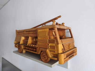 A wooden toy fire truck is displayed on a white shelf against a white wall. The fire truck is intricately carved and features various details such as a ladder, hoses, and wheels. The lighting suggests an indoor environment, possibly a home or a public space. The specific location in Vilnius, Lithuania, is not identifiable from the image.