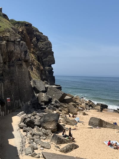 This is a medium-shot, eye-level photograph taken on a sunny day at Praia da Adraga, a beach located in Colares, Portugal. The image features a dramatic rocky cliff face on the left, with a path running along its base. The beach itself, covered in fine sand, is populated by several individuals engaged in various beach activities.

The cliff face is rugged and appears to be made of dark, layered rock, with some vegetation growing at its summit. A red and white warning sign is affixed to the lower portion of the cliff. At the base of the cliff, large boulders are scattered, leading down to the sandy beach.

The ocean occupies the right half of the frame, with calm blue water and gentle waves breaking near the shore. The horizon is visible in the distance, meeting a clear, pale blue sky.

On the beach, there are approximately 10-15 individuals. Some are sunbathing on towels, while others appear to be sitting or standing. Several groups are small, comprising 2-4 people. One individual is standing and stretching, and another is walking. There is a white umbrella on the sand, and a blue umbrella is visible in the distance. The people are dressed in casual beach attire, including swimwear. The overall atmosphere is one of leisure and relaxation on a summer day.