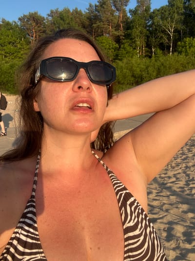 A woman wearing sunglasses and a zebra-print bikini top stands on a sandy beach, with trees in the background under a clear sky. The woman's arm is raised behind her head. The scene is bathed in the warm light of a setting sun, suggesting it is evening in Jūrmala, Latvia.