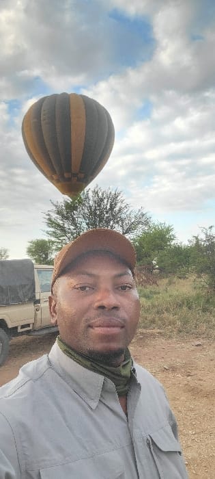 A dark-skinned male individual is captured in a vertical selfie in Seronera, Tanzania. He occupies the foreground, wearing a brown baseball cap, a grey long-sleeved collared shirt with two chest pockets, and an olive green neck gaiter. His gaze is directed forward towards the camera, maintaining a neutral expression. In the upper left background, a hot air balloon with vertical dark grey and tan stripes is visible, positioned aloft amidst a sparse treeline. To the man's left, a light-colored utility vehicle, likely a safari jeep, with a dark tarp covering its rear bed, is partially visible. The ground consists of reddish-brown dirt, and the sky is partly cloudy, displaying patches of blue between white and grey clouds.
