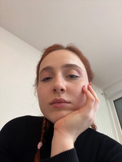 A young adult female with reddish-brown hair is depicted in a close-up, low-angle indoor shot, positioned in Eschau, France. She faces the camera directly, her head tilted slightly, with a neutral expression. Her right hand supports her chin and lower jaw; her fingernails exhibit red polish. She wears a black, ribbed long-sleeved top. A braid secured by a pink elastic tie is visible on her left side. The background consists of a plain, light-colored wall and ceiling, with the upper right portion of a window frame and vertical blinds partially visible on the right. A white bracket or fixture is mounted on the wall near the window.
