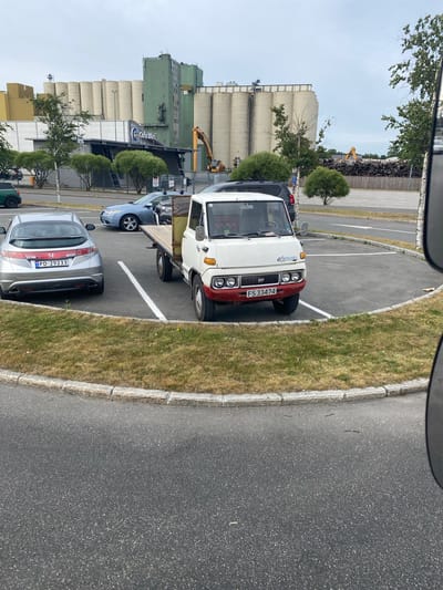 This is a vertically oriented photograph taken from a slightly elevated perspective, possibly from inside a vehicle, looking down at a parking lot and an industrial area. The scene is likely set in Larvik, Norway, given the provided location context.
The primary subjects are several vehicles parked in a lot. In the foreground, a silver hatchback car with a license plate reading "PD 29233" is prominently visible. Behind it, a dark-colored car, partially obscured, is also parked. Further back, a white and red flatbed truck with the license plate "FS 33474" is parked. The truck appears to be older.
The setting includes a paved parking lot with white painted lines demarcating parking spaces. To the right of the parking lot, there is a strip of dry, yellowing grass bordered by a concrete curb. In the background, a large industrial complex dominates the scene. This complex features several tall, cylindrical silos, some of which are light beige and others are greenish. Adjacent to the silos are multi-story buildings with a green facade. An excavator is visible in the distance, positioned near the industrial buildings, suggesting ongoing construction or demolition work. Trees with green foliage are scattered throughout the scene, adding some natural elements to the industrial landscape. The sky is overcast and pale, indicating a cloudy day, possibly with mild weather. There are no people visible in the photograph.
The overall impression is one of a functional, industrial environment on a typical, overcast day. The presence of parked vehicles suggests a working day, but the lack of people in the frame creates a sense of stillness.