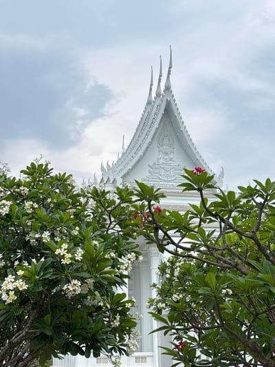 The image captures a low-angle view of a white, ornately decorated roofline, possibly belonging to a Buddhist temple, situated in Muang Pattaya, Thailand. The sky is overcast with grey clouds. Lush green trees with white and pink blossoms are partially obscuring the building and filling much of the foreground, their branches extending into the frame. The roofline's decorative elements include tiered, sharp pointed accents and a central embossed design. The building itself is white, while the overall setting includes vegetation and a cloudy sky.