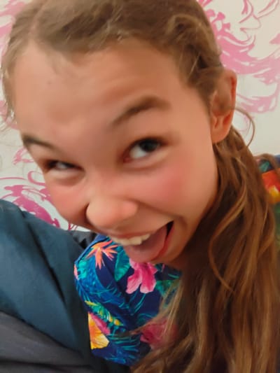 The image is a close-up, candid selfie of a young girl with light brown hair and fair skin. She is making a playful, exaggerated facial expression with her eyes squinted and her mouth open, sticking her tongue out slightly. She is wearing a colorful, tropical-print shirt with flowers and leaves. The background appears to be a wall with a pink floral pattern. The girl is partially leaning against a dark blue fabric, possibly a blanket or piece of clothing. The lighting is soft and appears to be from an indoor source. There are no specific identifiable landmarks or features that would indicate a location in Podsteрки, Russia.