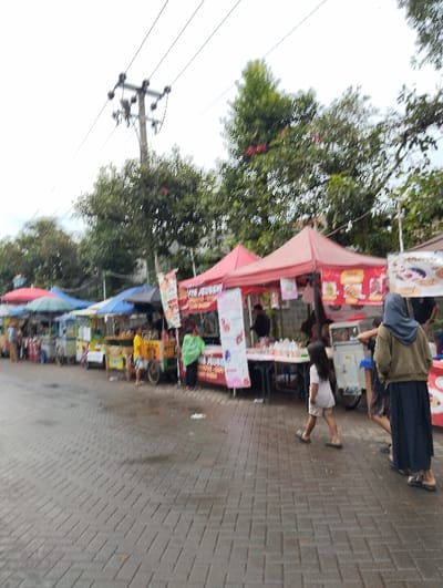 The scene depicts a street food market in Banjaran, Indonesia, located at coordinates -7.047 latitude and 107.58 longitude. The market is comprised of several stalls, each with a red canopy. There are various individuals interacting with the stalls, including vendors, shoppers, and onlookers. The vendors are preparing and displaying food items for sale. The shoppers are browsing the food selection and interacting with the vendors. Some individuals are seated at tables while others are standing or walking. There are various food items displayed for sale, including snacks, drinks, and cooked meals. The market is located on a paved street. The weather is overcast. The street is wet, suggesting that it may have recently rained. The scene is bustling with activity. Trees can be seen in the background, providing shade for the market. Several electrical wires are visible overhead.