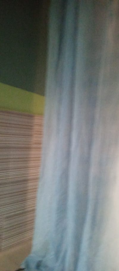 This is a blurry, abstract image with a focus on colors and textures. On the left side of the frame, there are horizontal lines in shades of brown and beige, suggesting a textured surface like wood paneling or blinds. Above this textured area, there is a horizontal band of muted green, and above that, a darker, almost black area. The right two-thirds of the image are dominated by a large, soft expanse of light blue, which appears to be a curtain or fabric. The light blue has vertical folds and wrinkles, creating a sense of depth and texture. The lighting is subdued, with some areas brighter than others, and the overall impression is one of diffused light. There is no clear indication of people, specific objects, or a distinct activity taking place. The scene appears to be indoors, possibly a room with a window covered by a light blue curtain. The context provided, Zanzi, Nigeria, does not offer specific visual cues within the image itself. The blurriness of the image obscures any fine details, making it difficult to determine the time of day, weather, or specific emotions conveyed. There is no visible text in the image.