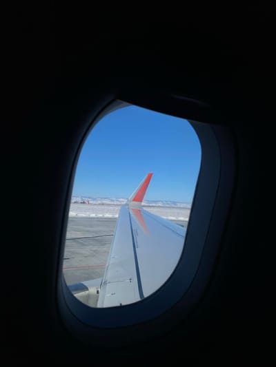 Through an airplane window, a view of the clear blue sky above the plane's wing is visible. The wing features a striking orange winglet. Below, a ground surface, possibly an airport tarmac, appears alongside a snowy landscape. The interior of the plane is dark, contrasting with the bright daylight outside the window.
